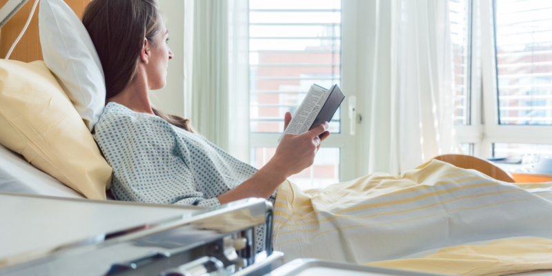 A woman in a hospital bed reading a bible and looking out the window.