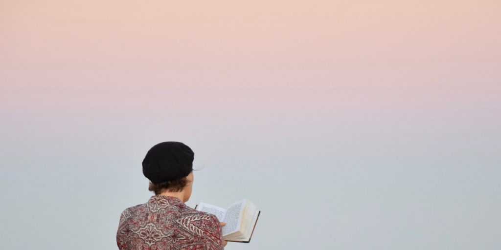 a photo of a woman in a black hat reading the old testament against a pink sunset